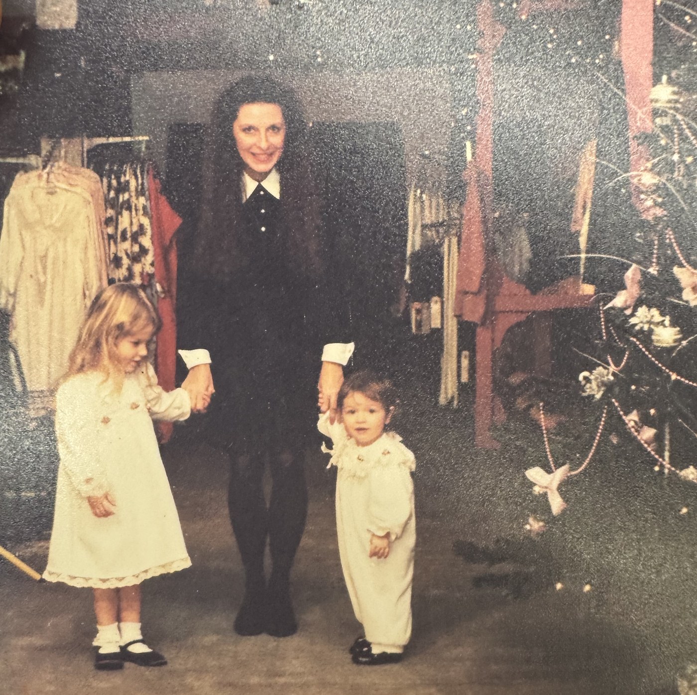 Two small girls in white dresses holding hands with a woman in black inside the shop, garment racks behind them and a Christmas tree to the right