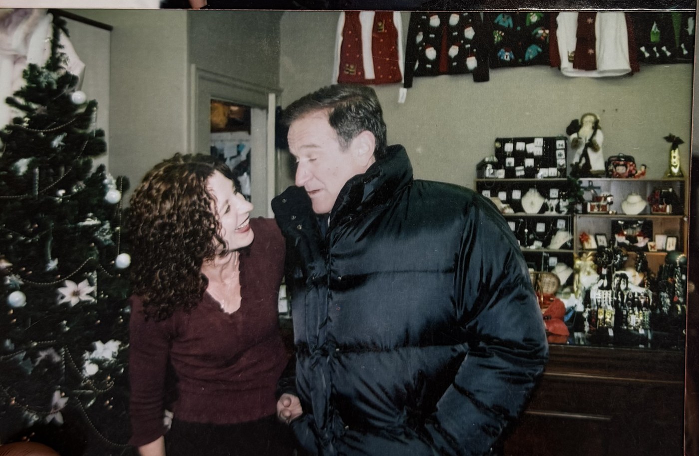 Robin Williams laughing with a woman inside Amarettos, beside a decorated Christmas tree, with Christmas sweaters on the wall and gift cabinets behind them